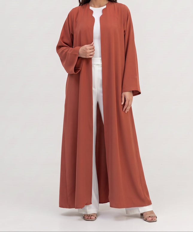 A woman modeling a long, flowing terracotta-colored abaya over a white jumpsuit, standing against a plain light gray studio background.